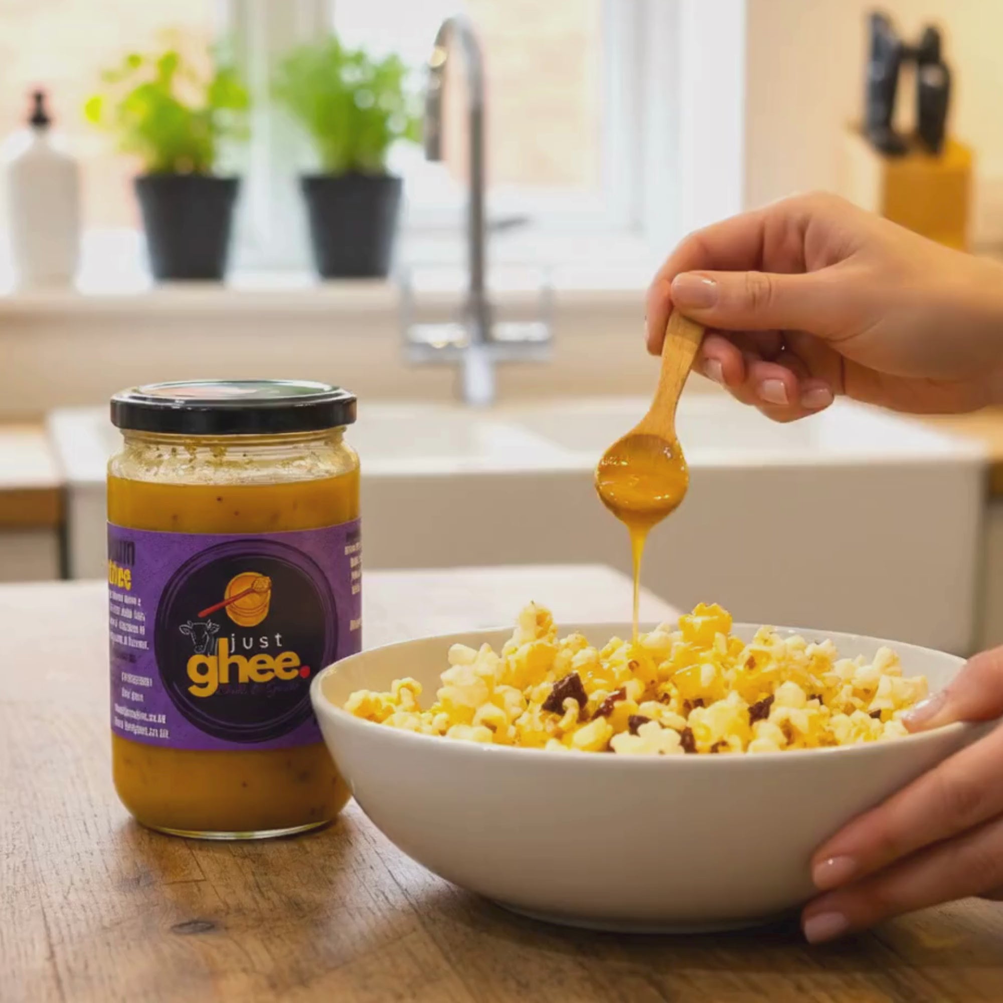 Load video: A close-up scene in a bright kitchen shows a jar of Just Ghee placed on a wooden countertop next to a white bowl of freshly popped popcorn. A person holds a small wooden spoon above the bowl, slowly drizzling warm, golden ghee over the popcorn. The ghee flows smoothly in a thin stream, coating the popcorn and giving it a glossy, rich appearance. In the softly blurred background, a kitchen sink and small green plants sit by a sunlit window, creating a warm, homely atmosphere.